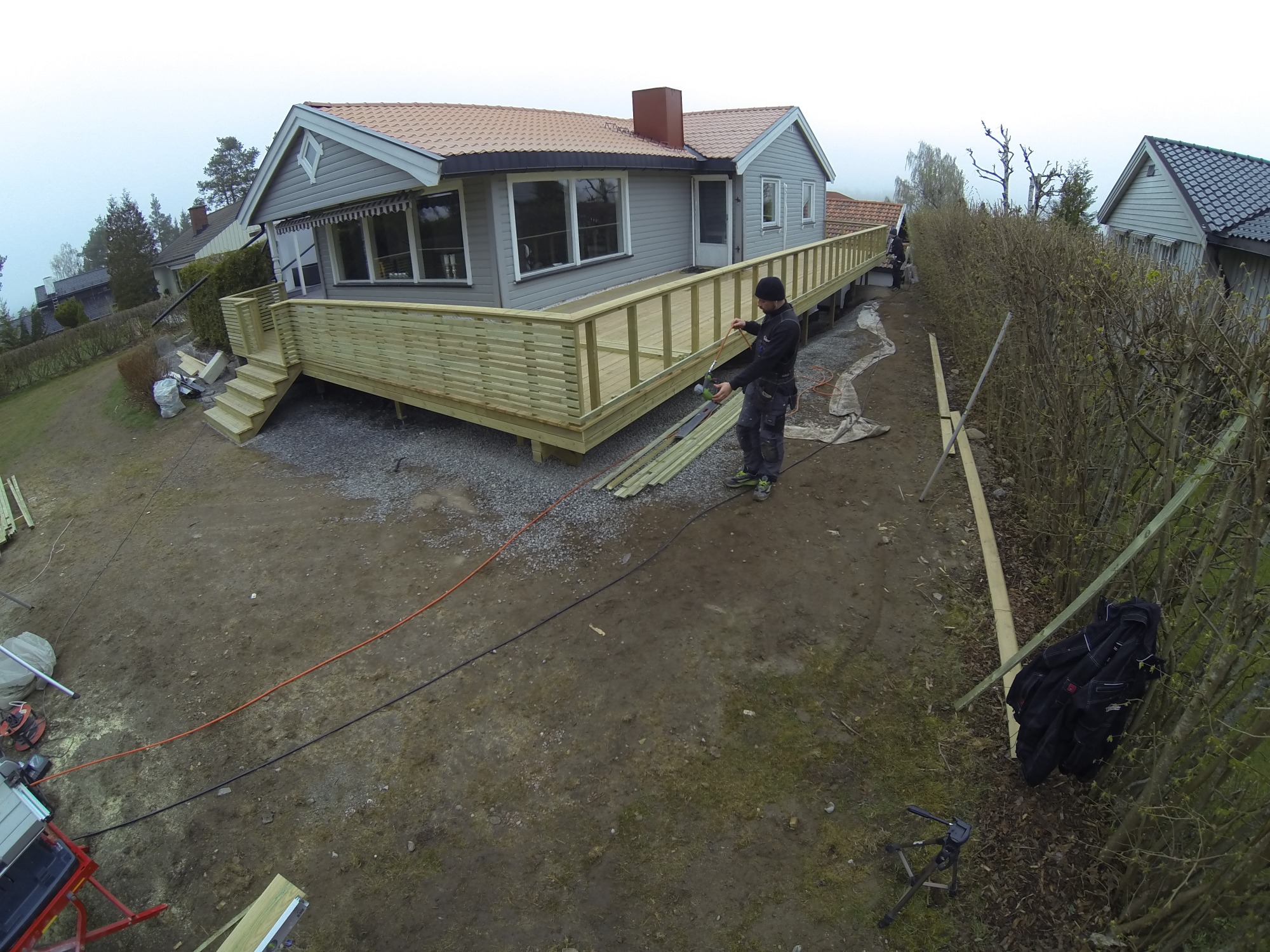 Montaż drewnianego tarasu przy szarym domu z czerwonym dachem, widoczne narzędzia i pracownik w czapce podczas pracy, kabel elektryczny na ziemi.