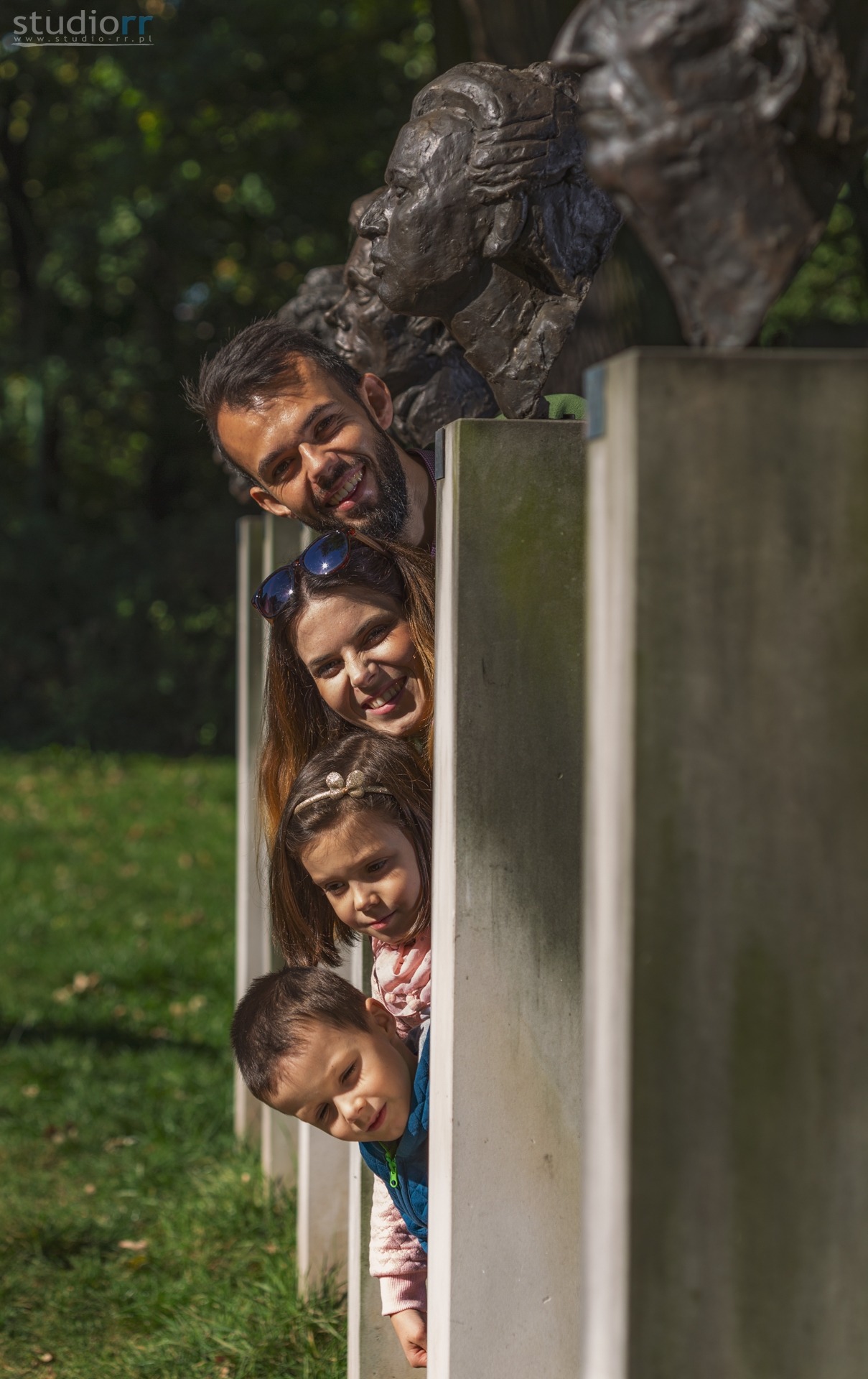 Uśmiechnięta rodzina, tata, mama, córka z kokardą i syn, wyglądają zza betonowych słupków, w tle rzeźba plenerowa i zieleń parku.