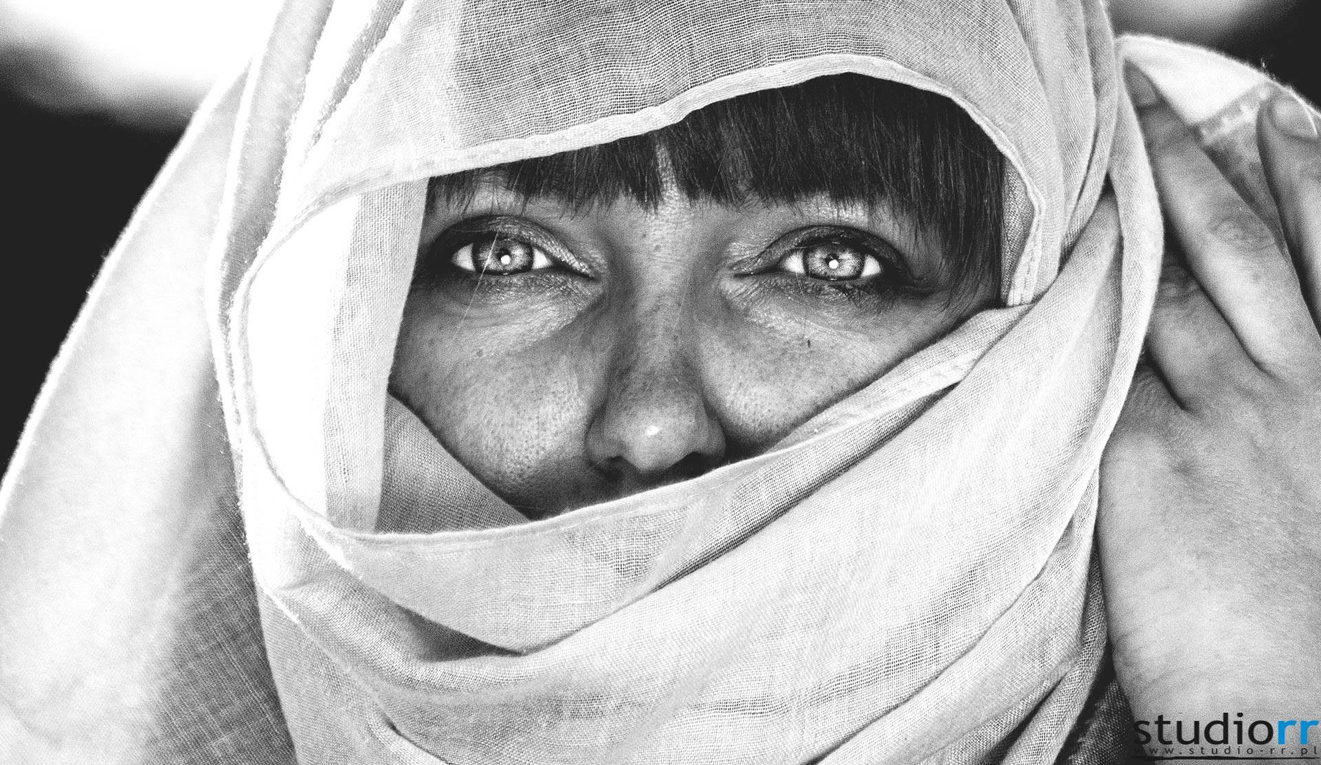 Czarno-biały portret kobiety z intensywnym spojrzeniem, owiniętej w delikatną, wzorzystą tkaninę zakrywającą usta i nos, z widocznymi zmarszczkami wokół oczu.