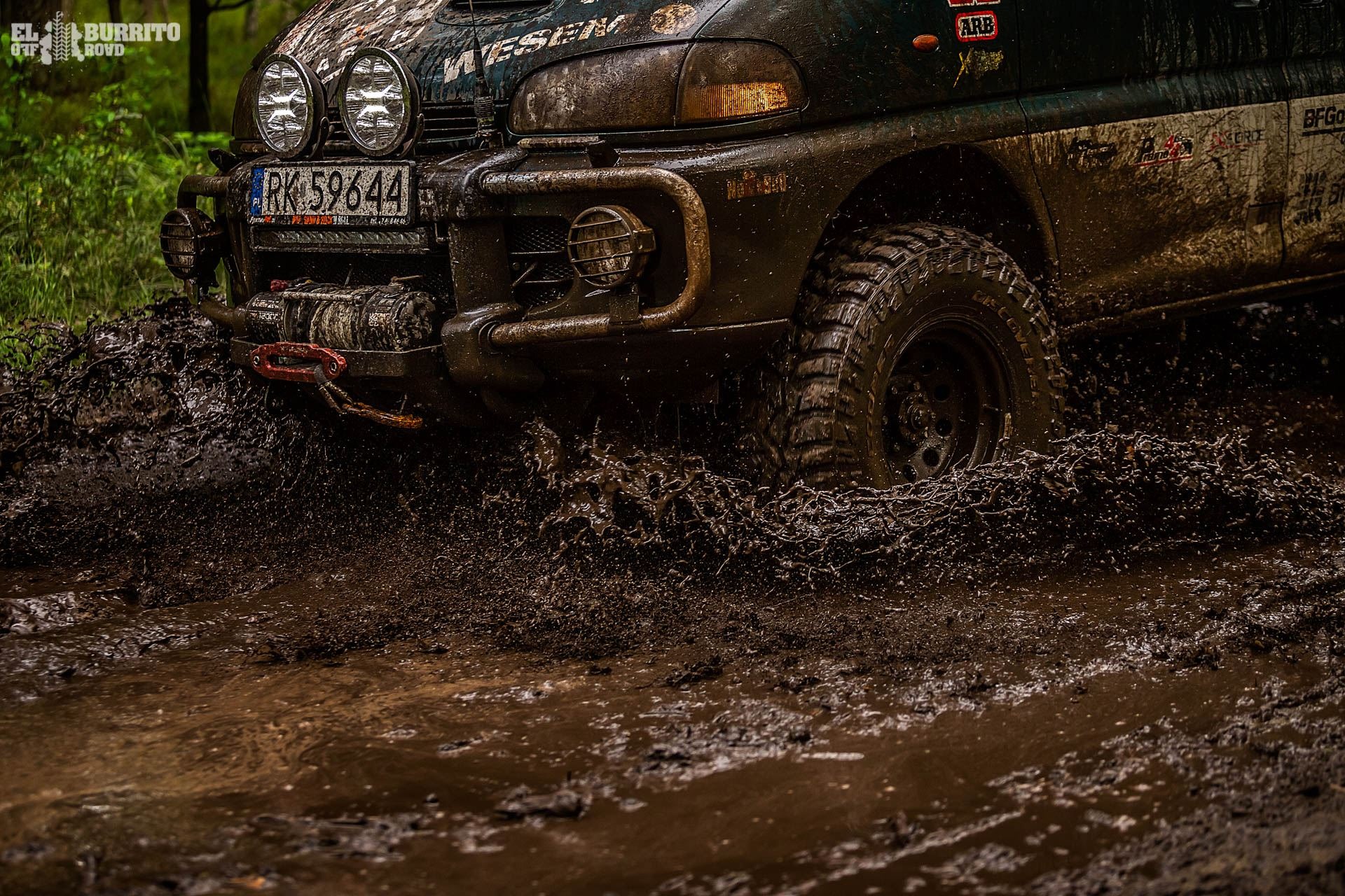 Zielony samochód terenowy z wciągarką i dodatkowymi reflektorami przedziera się przez błotnistą wodę, rozbryzgując ją na boki.