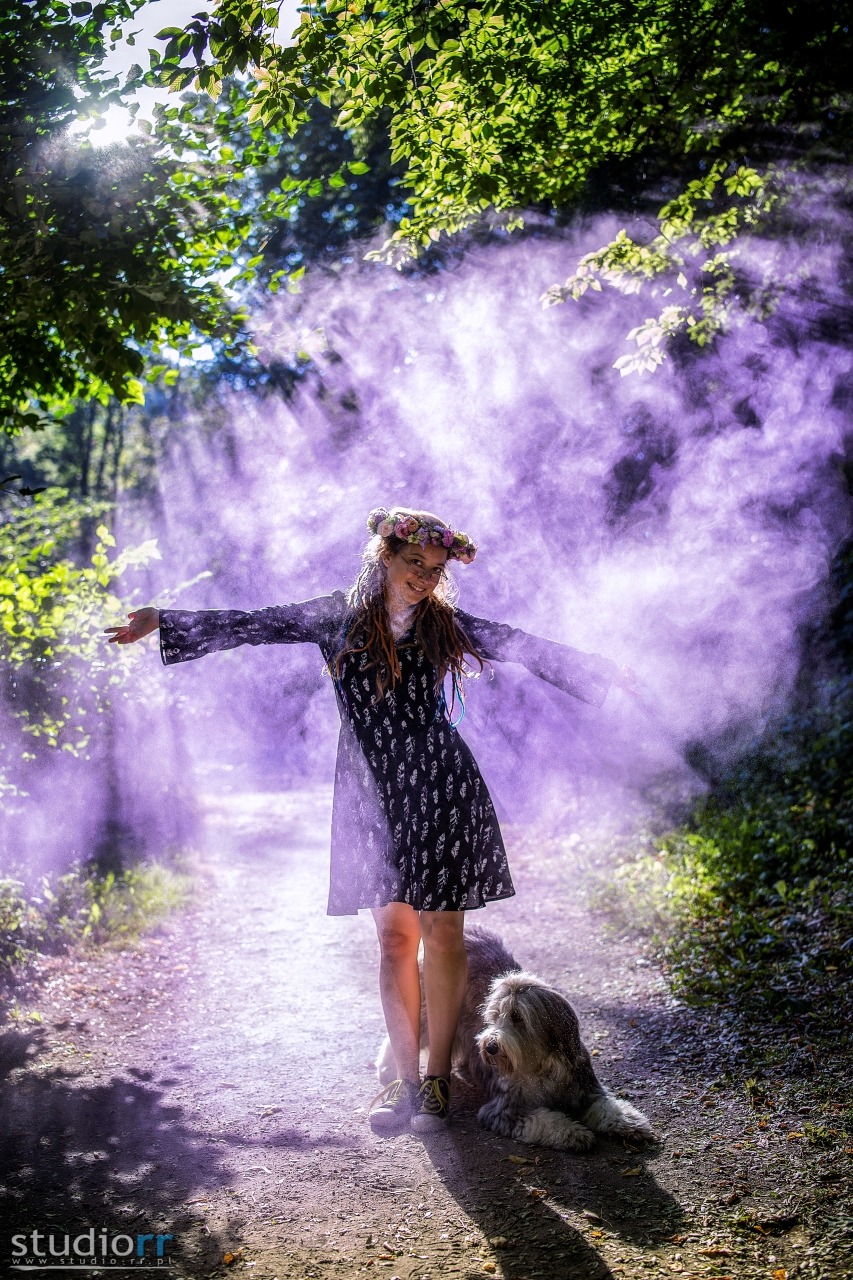 Młoda kobieta w kwiecistym wianku i sukience z rozpostartymi ramionami, otoczona fioletową mgłą na leśnej ścieżce, pies leżący u jej stóp.