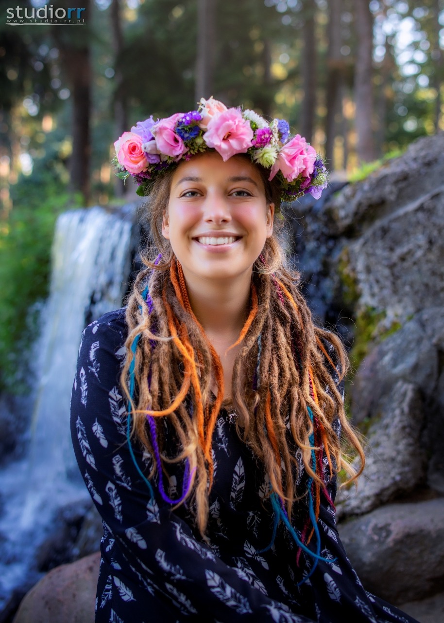 Uśmiechnięta młoda kobieta z dredami ozdobionymi kolorowymi pasemkami i wiankiem z różowych i fioletowych kwiatów na głowie, na tle skalnego wodospadu i drzew.