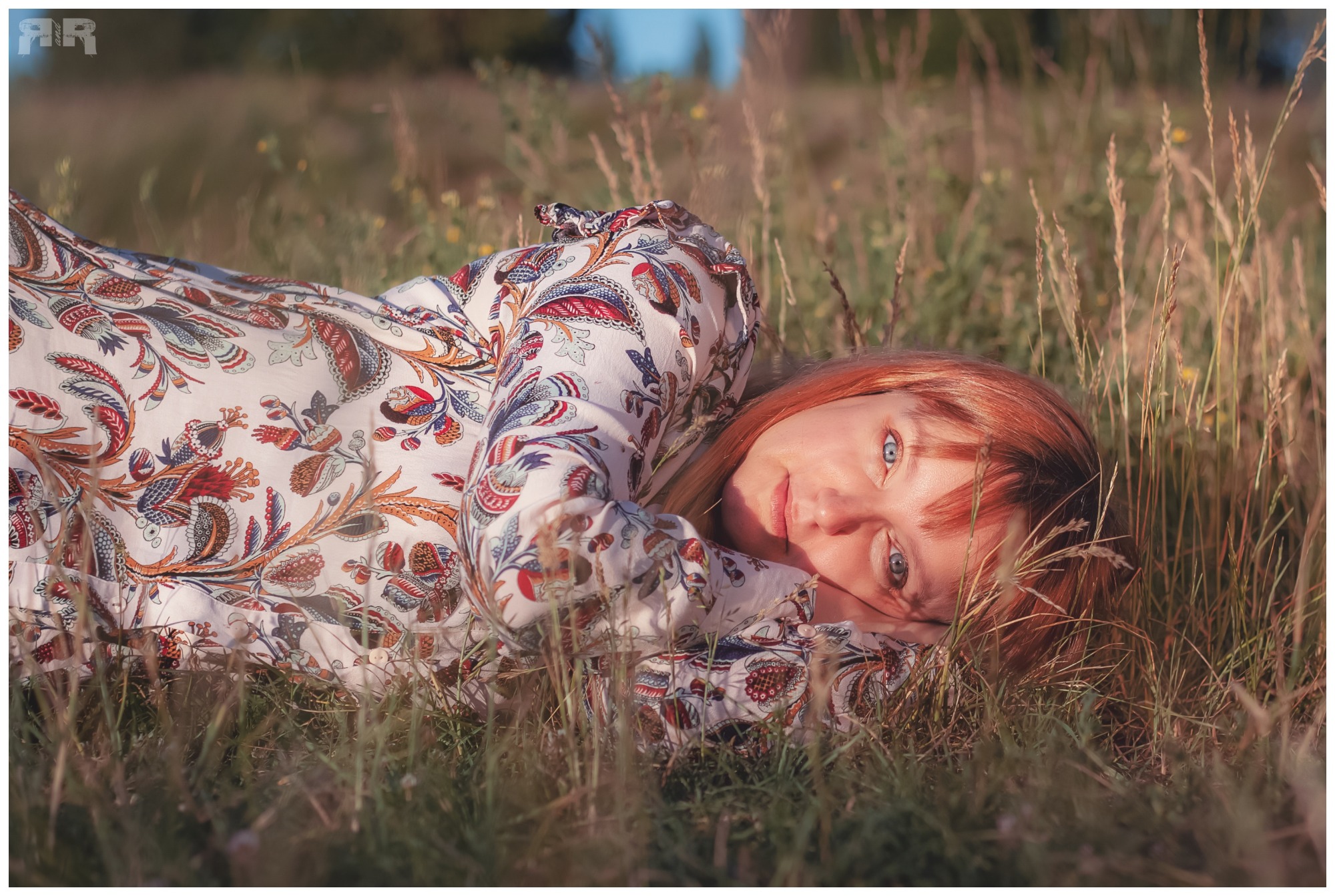 Kobieta z rudymi włosami i niebieskimi oczami leży na łące w kwiecistej sukience, patrząc w obiektyw.