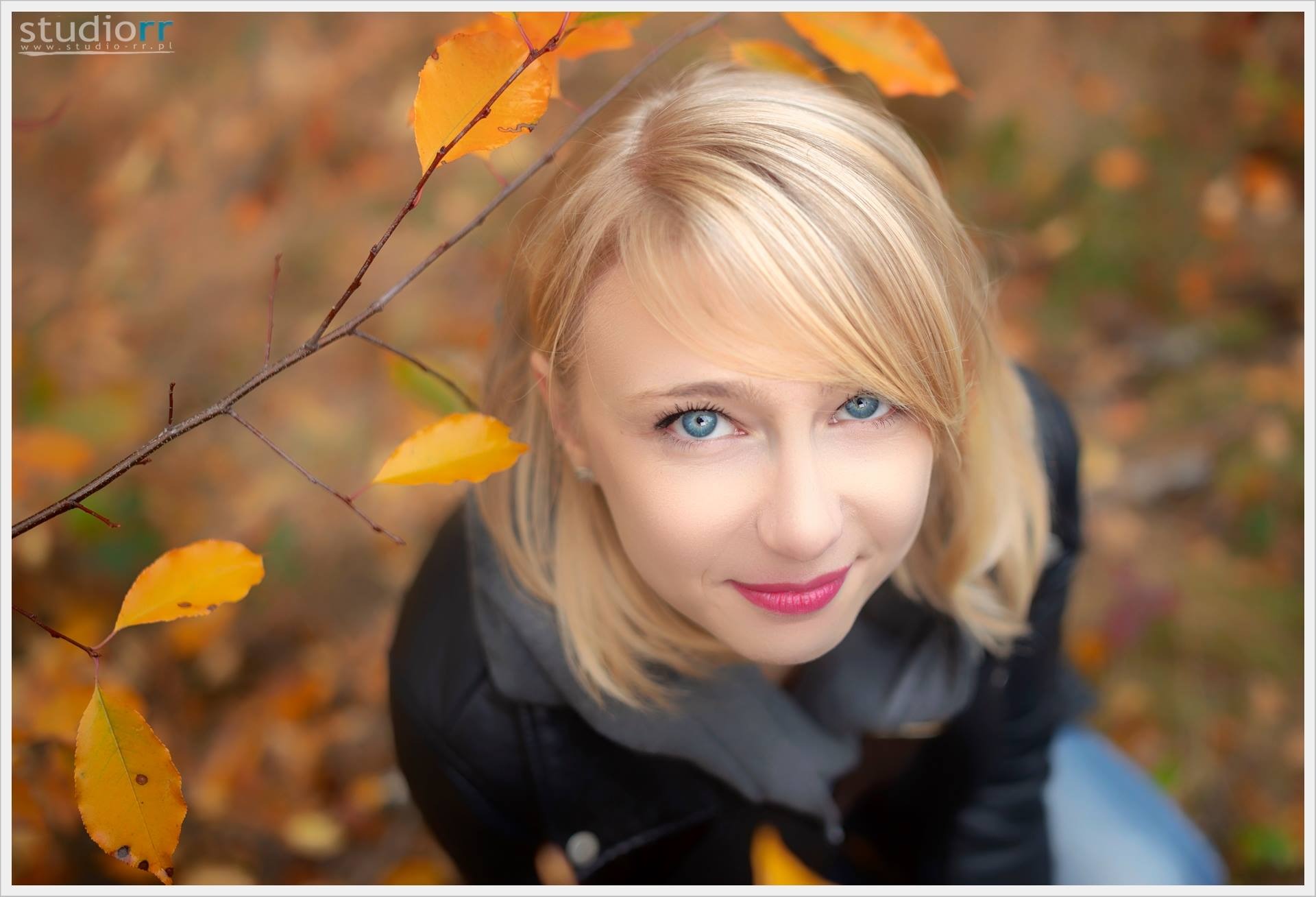 Portret uśmiechniętej blondynki z niebieskimi oczami, spoglądającej w górę, z jesiennymi liśćmi w tle i gałązką z liśćmi w lewym górnym rogu kadru.