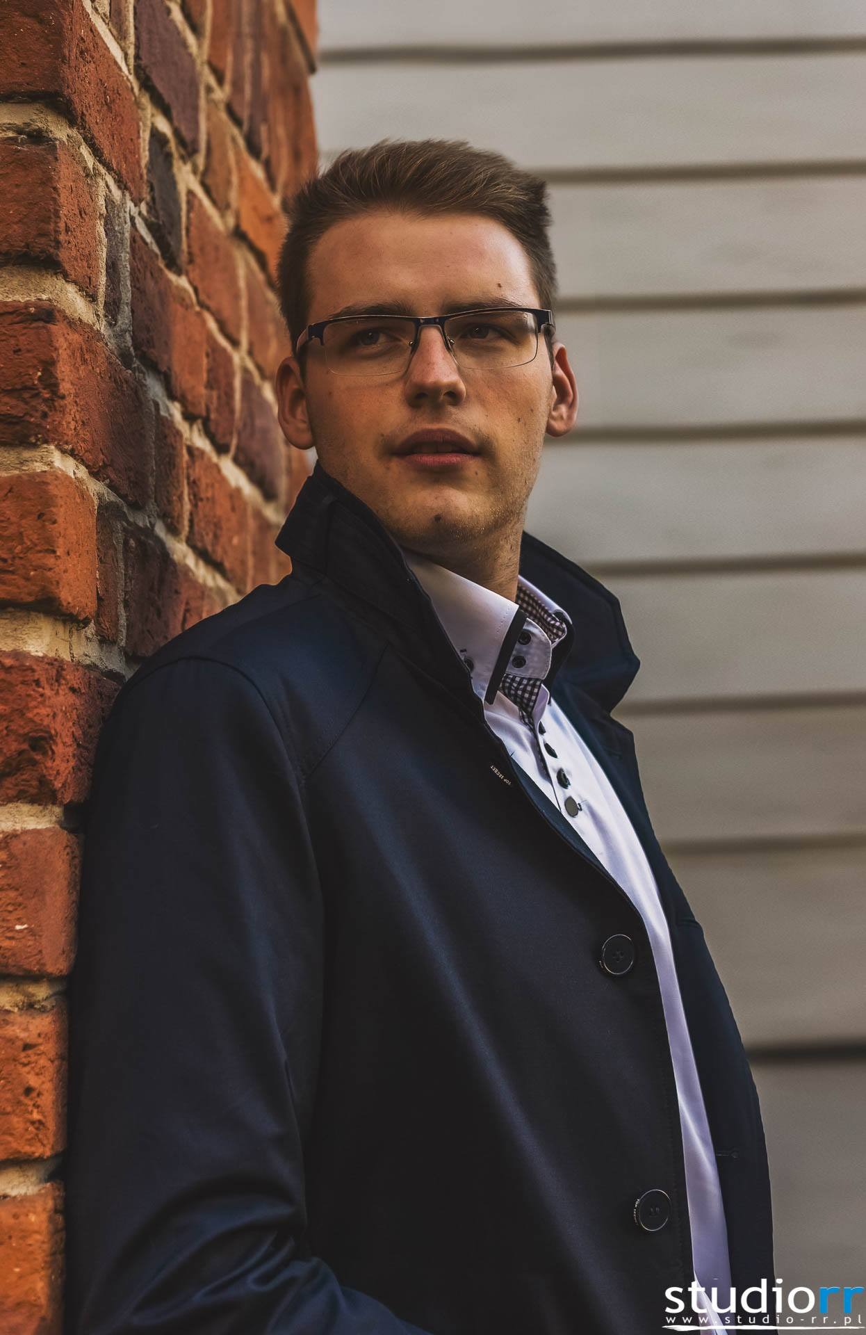 Portret młodego mężczyzny w okularach, opierającego się o ceglaną ścianę w ciemnym płaszczu i białej koszuli z wzorzystym kołnierzykiem, spoglądającego w górę.