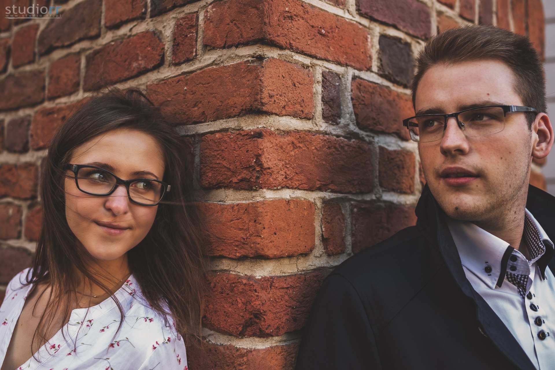 Młoda para w okularach, oparta o ceglany mur. Kobieta w kwiecistej bluzce spogląda w lewo, mężczyzna w garniturze patrzy w prawo.