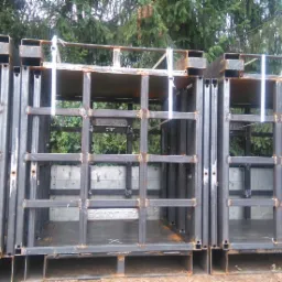 Seryjnie ustawione, spawane, metalowe ramy z kratownicą, z widocznymi śladami spawania, niektóre z naklejkami, ułożone na paletach, z zielonym tłem drzew