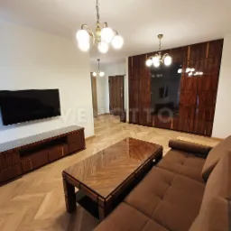 Salon z brązową sofą, stolikiem kawowym z fornirem, szafą wnękową z ciemnego drewna i telewizorem na ścianie, oświetlony lampami w stylu retro.