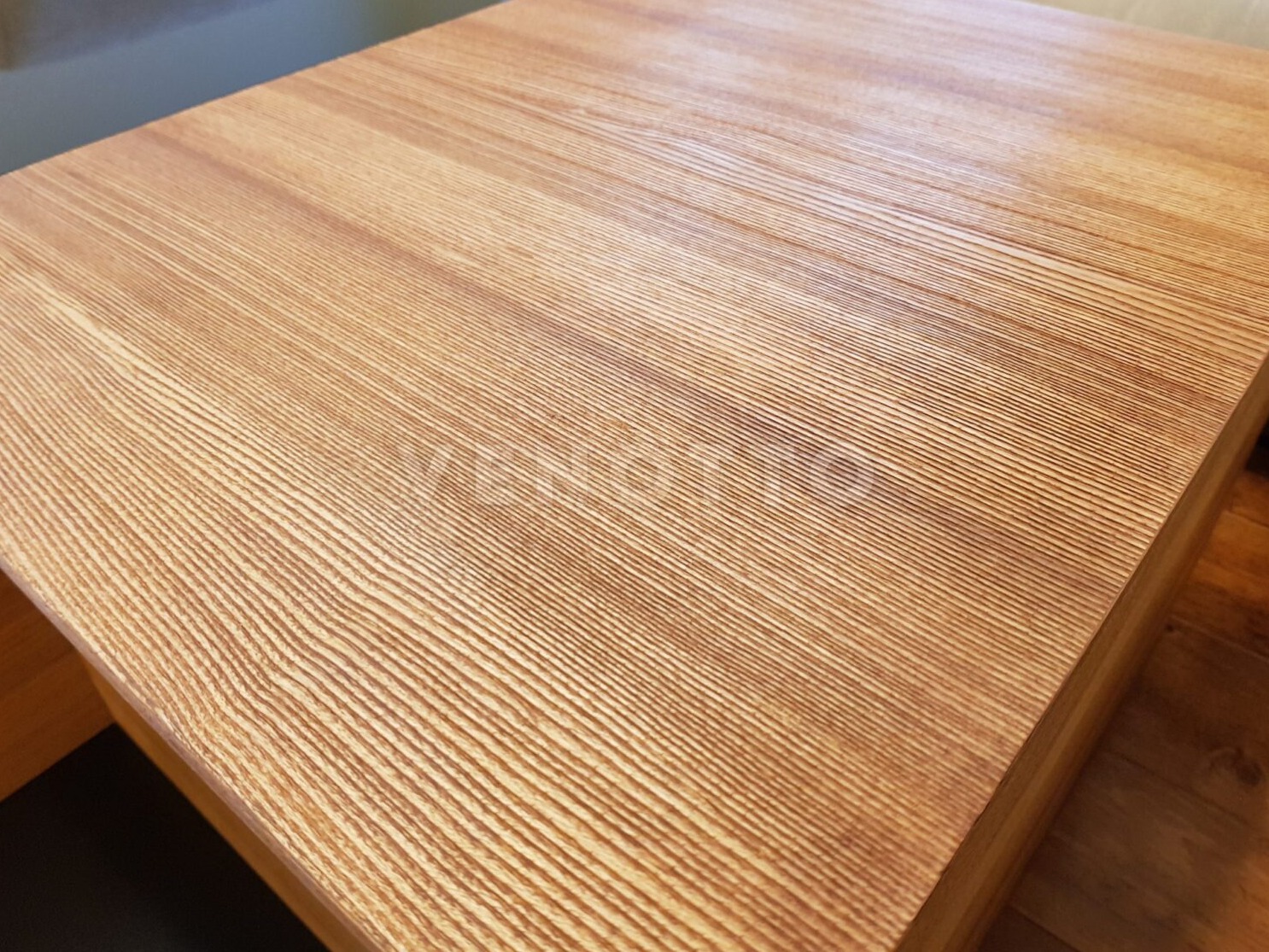 Jasny blat drewniany z wyraźnym usłojeniem, widok z góry pod kątem, ukazujący krawędź blatu i fragment podłogi.
