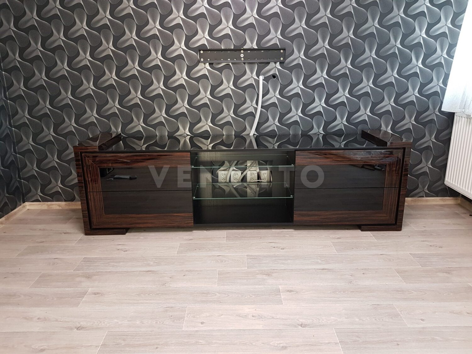 Długa, niska komoda RTV z ciemnego drewna z błyszczącymi czarnymi frontami szuflad i szklaną półką, na tle wzorzystej tapety i jasnej podłogi.