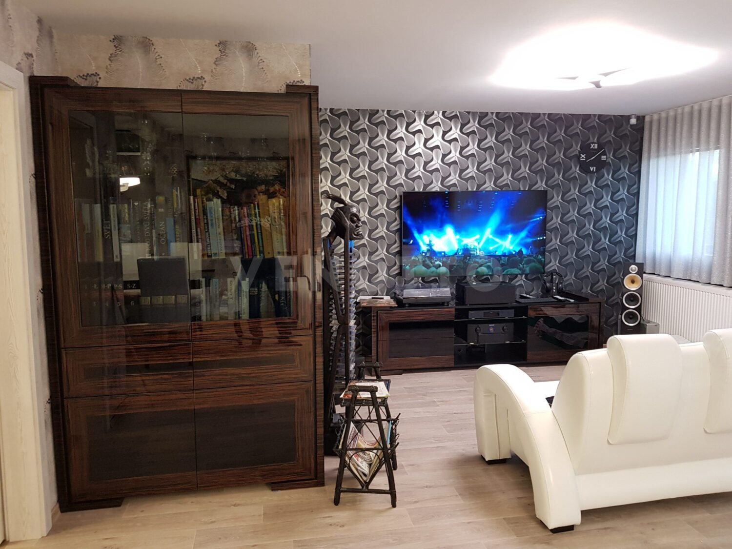 Salon z dużą, przeszkloną biblioteczką z ciemnego drewna, telewizorem na ścianie z wzorzystą tapetą i białą sofą.