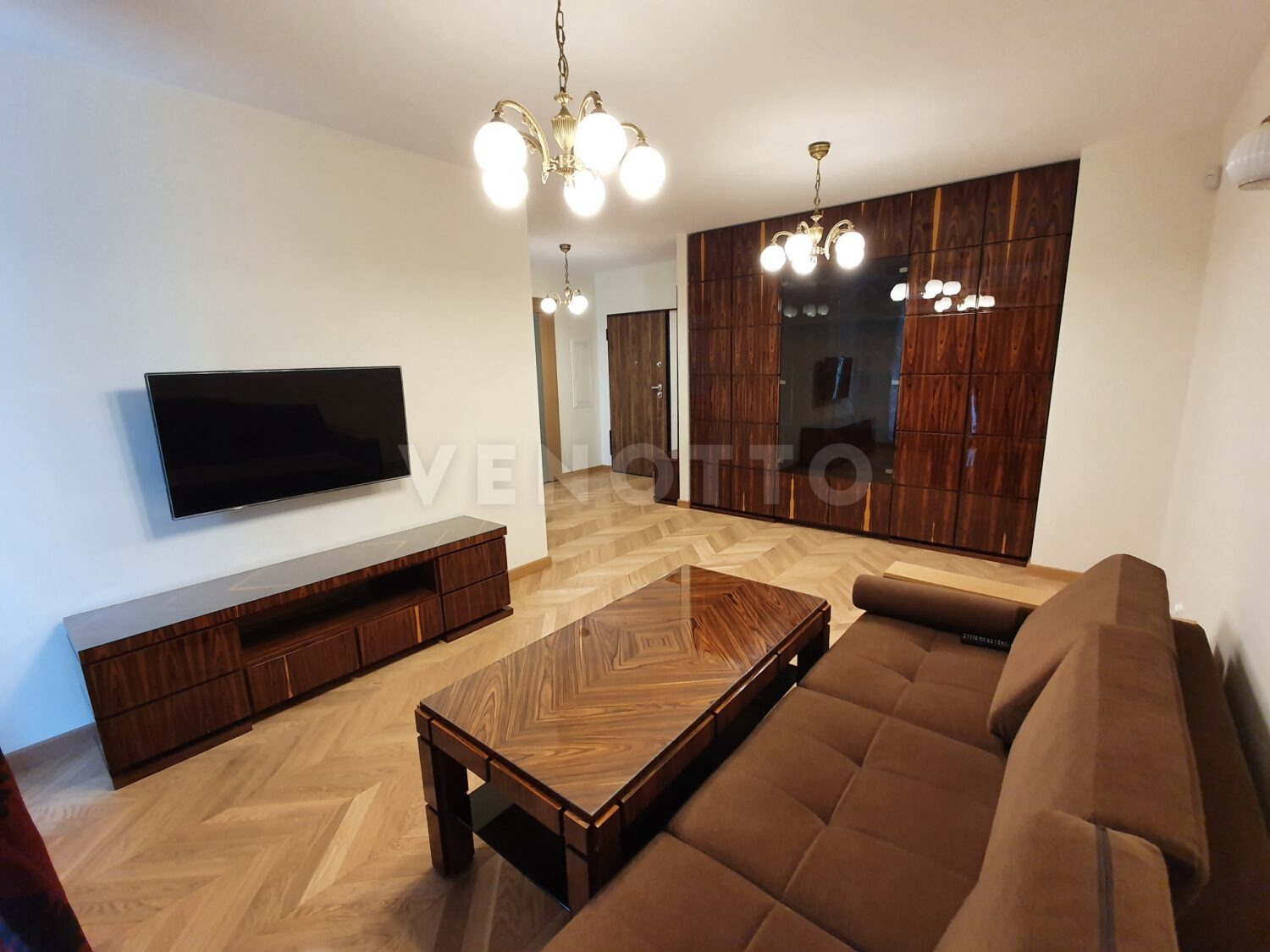 Salon z brązową sofą, stolikiem kawowym z fornirem, szafą wnękową z ciemnego drewna i telewizorem na ścianie, oświetlony lampami w stylu retro.