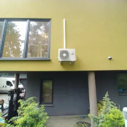 Zewnętrzna jednostka klimatyzacji Mitsubishi zamontowana na elewacji domu o nietypowym, dwukolorowym wykończeniu, z widocznymi elementami instalacyjnymi i otoczeniem posesji.