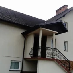 Dwupiętrowy dom z jasną elewacją, ciemnym dachem z blachy falistej i balkonem z ozdobną, czarną balustradą. Widoczny fragment ogrodu i komin z cegły.