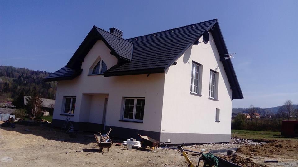 Nowo wybudowany dom jednorodzinny z ciemną dachówką i jasną elewacją, widoczne narzędzia budowlane i teren w trakcie zagospodarowywania.