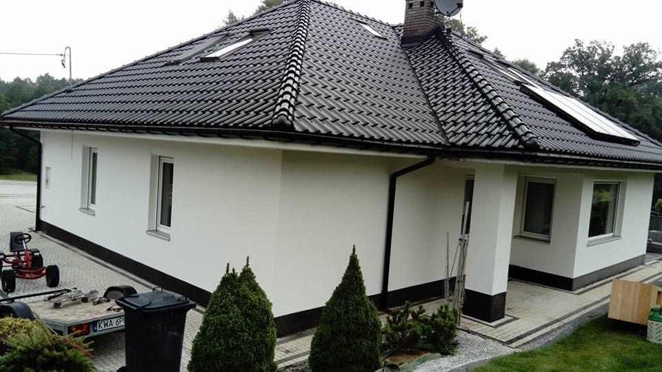 Nowoczesny dom jednorodzinny z czarną dachówką karpiówką, białymi ścianami i ciemną podmurówką, widoczne okna dachowe i komin, trawnik i ozdobne iglaki przed budynkiem.