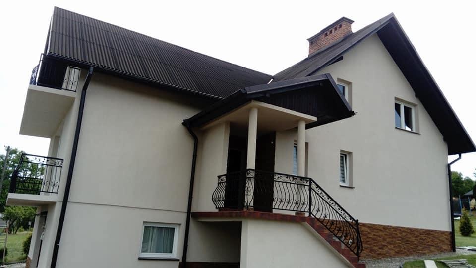 Dwupiętrowy dom z jasną elewacją, ciemnym dachem z blachy falistej i balkonem z ozdobną, czarną balustradą. Widoczny fragment ogrodu i komin z cegły.