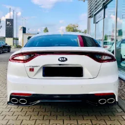 Biały samochód marki Kia Stinger z widocznym logo i czterema chromowanymi końcówkami wydechu, zaparkowany na kostce brukowej przed salonem samochodowym z dużymi szklanymi witrynami.