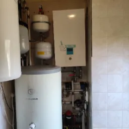 Kotłownia z białym bojlerem marki Biawar, białym zbiornikiem wyrównawczym i wiszącym kotłem gazowym Aquarea, widoczne liczne rury i zawory.