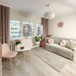 Jasny salon z sofą, dywanem, stolikami, krzesłem i roślinami w doniczkach, utrzymany w pastelowej kolorystyce z przewagą różu i szarości, z widokiem na blok mieszkalny za oknem.