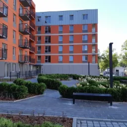 Nowoczesny budynek mieszkalny z pomarańczową elewacją i czarnymi balkonami, otoczony zadbanym terenem zielonym z krzewami, kwiatami i brukowanym chodnikiem.