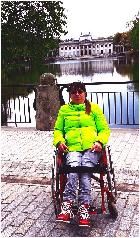 Kobieta w neonowej kurtce i czerwonych butach siedzi na wózku inwalidzkim na tle Pałacu na Wodzie w Łazienkach Królewskich w Warszawie.