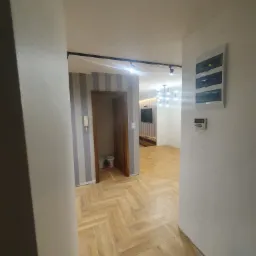 Korytarz z parkietem w jodełkę, widok na salon z oświetleniem punktowym i dekoracyjnym żyrandolem, ściany w odcieniach bieli i szarości, puszki farb w tle.