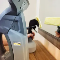 Szary odkurzacz do okien Kärcher Professional i spryskiwacz z żółtą nakładką z mikrofibry, odbijające się w lustrze na drewnianej komodzie.