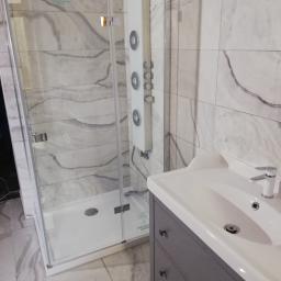 Majster - Nowoczesna kabina prysznicowa z panelem natryskowym i umywalką z szafką w łazience wykończonej płytkami imitującymi marmur.