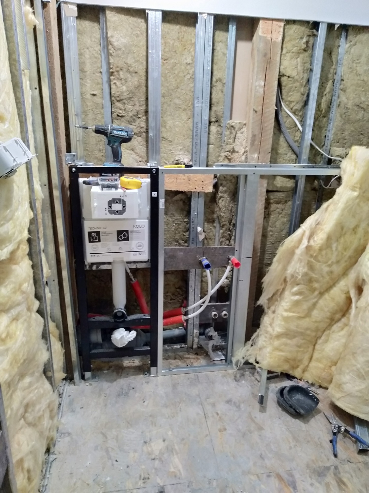 Stelaż podtynkowy WC marki Koło zamontowany w trakcie prac wykończeniowych łazienki, widoczne izolacja z wełny mineralnej, elementy instalacji wodnej z zaworami, wiertarka akumulatorowa na stelażu.