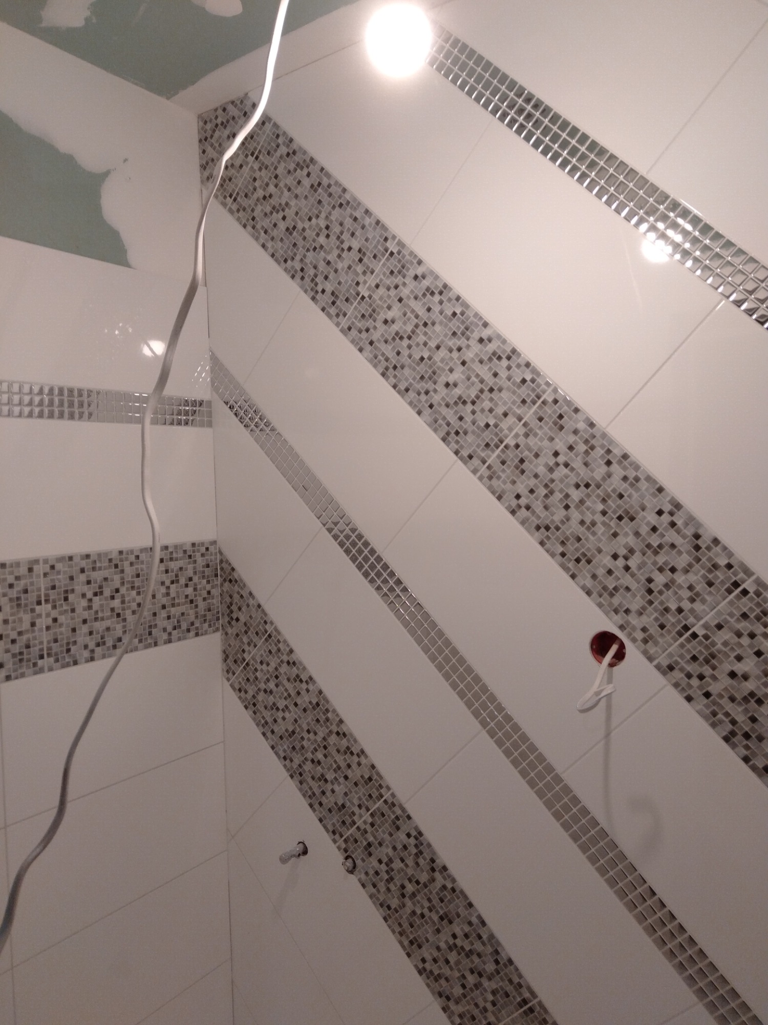 Ukończona łazienka z białymi płytkami ułożonymi ukośnie, ozdobnymi pasami z mozaiki, widocznym gniazdkiem elektrycznym i wystającym kablem, sufit częściowo przygotowany do malowania.