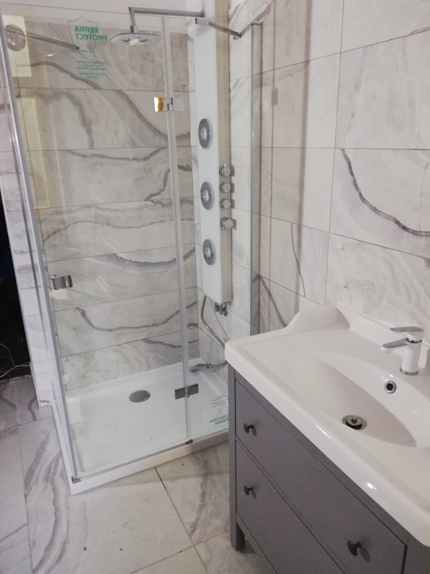 Nowoczesna kabina prysznicowa z panelem natryskowym i umywalką z szafką w łazience wykończonej płytkami imitującymi marmur.