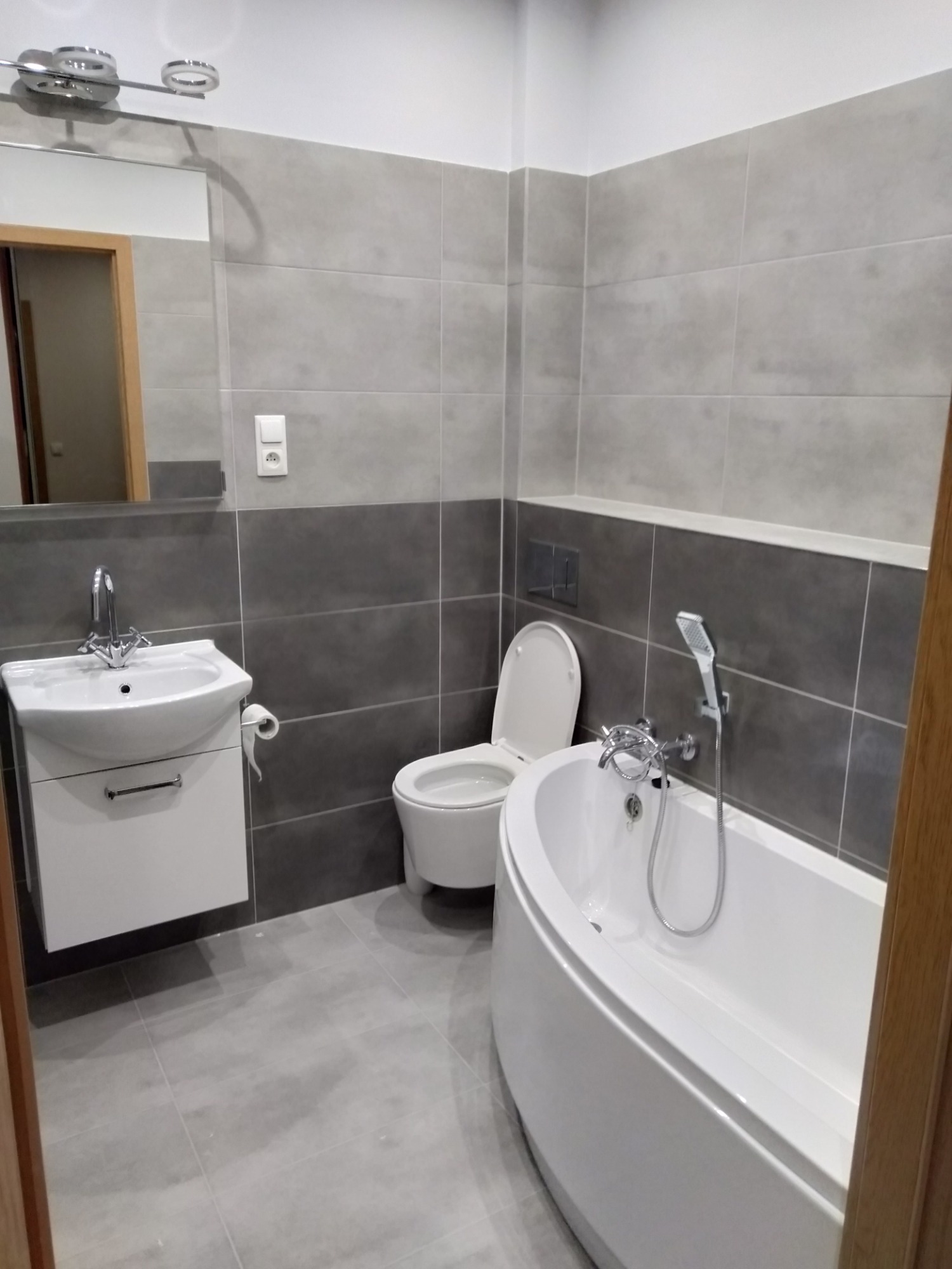 Wykończona łazienka z szarymi płytkami, białą armaturą i podwieszaną toaletą, widok z wejścia.