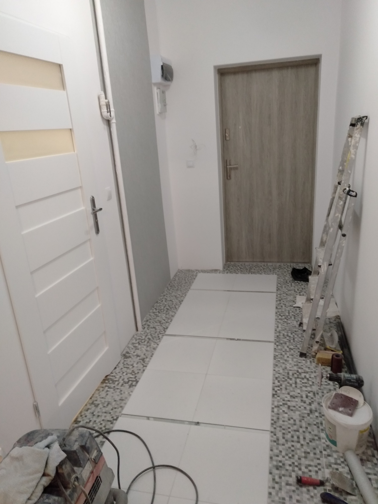 Korytarz w trakcie układania białych płytek podłogowych na mozaikowej wylewce, widoczna drabina, narzędzia i sprzęt budowlany.