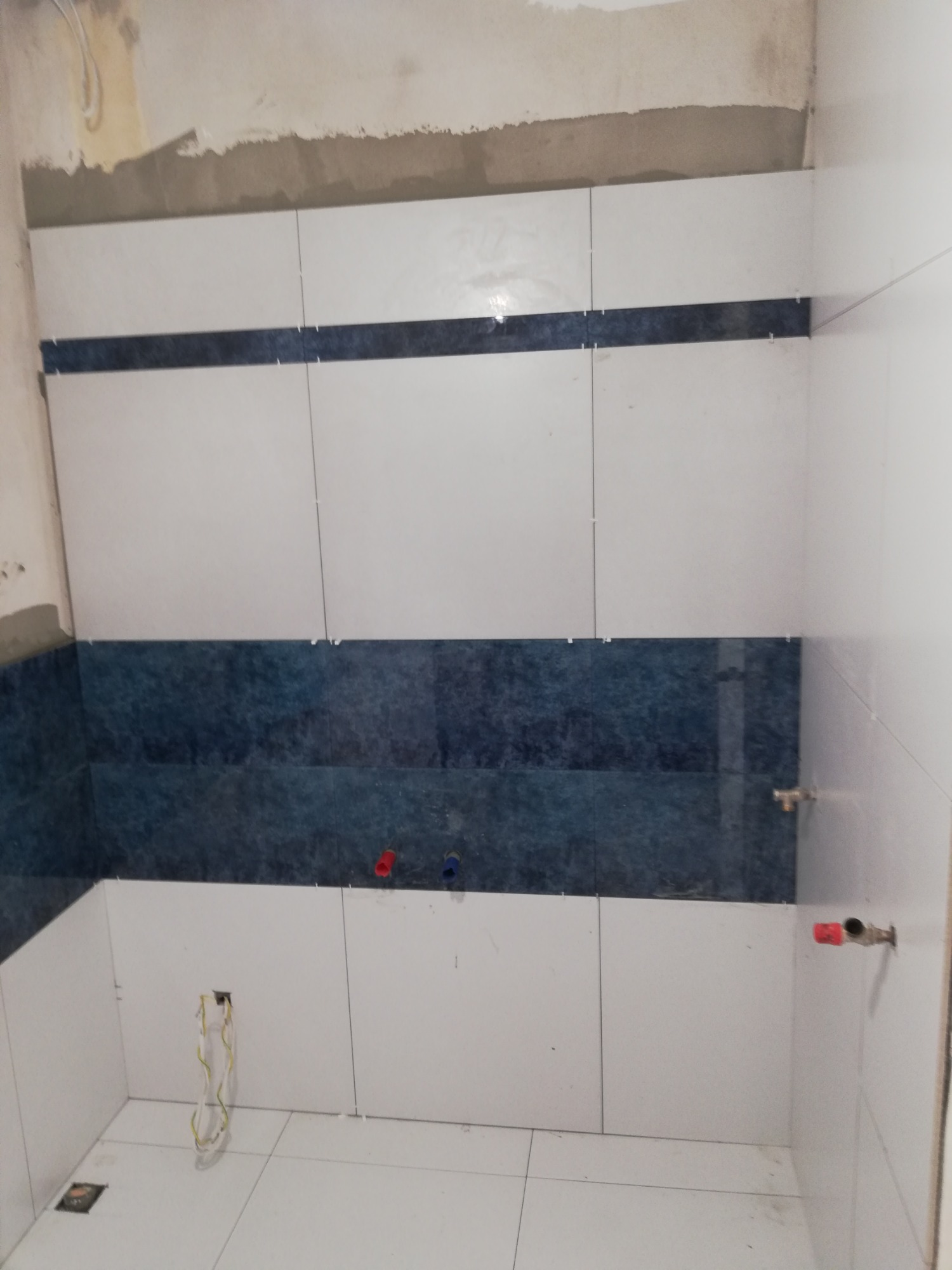 Wnętrze łazienki w trakcie prac wykończeniowych: ułożone białe i niebieskie płytki na ścianach, widoczne wystające rury instalacji wodnej oraz kable elektryczne ze ściany, fugi zabezpieczone...