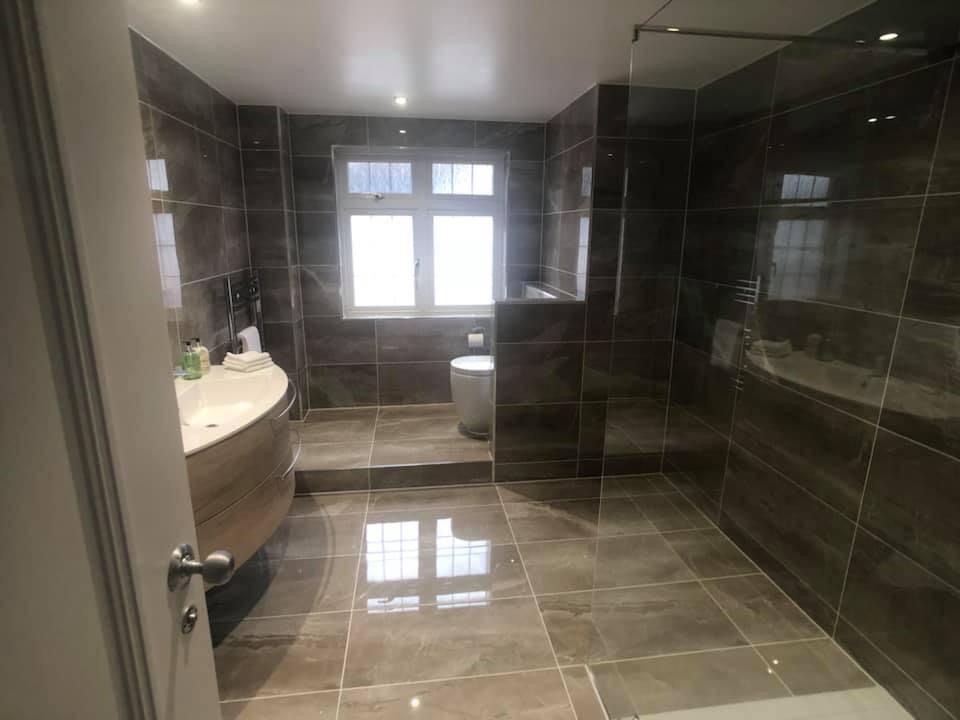 Nowoczesna łazienka z brązowymi, marmurkowymi płytkami, owalną umywalką z drewnianą szafką, toaletą na podwyższeniu i kabiną prysznicową ze szklaną ścianką.