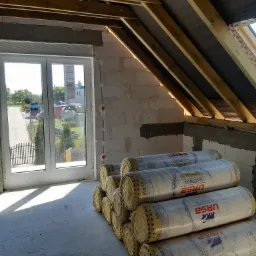 Stos rolek wełny mineralnej Ursa Glasswool Gold 35 na poddaszu w trakcie prac izolacyjnych, widoczne krokwie dachu i okno z widokiem na zewnątrz.