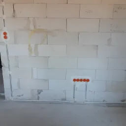 Surowa ściana z betonu komórkowego z widocznymi puszkami instalacyjnymi i przewodami elektrycznymi, częściowo zamurowanymi, z odręcznymi notatkami na ścianie.