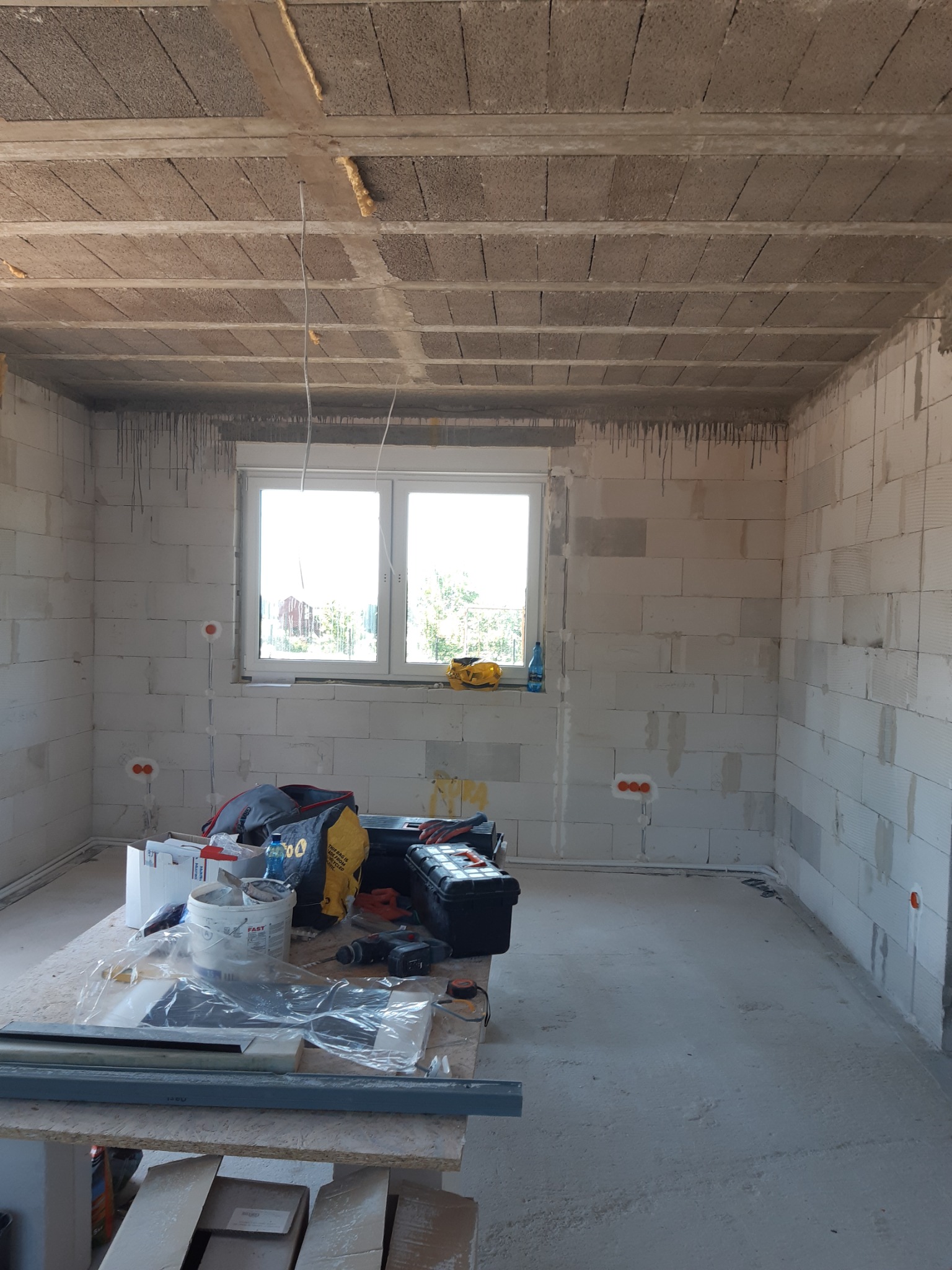 Surowy stan wnętrza z bloczków, widoczne puszki instalacyjne, narzędzia i materiały na prowizorycznym stole warsztatowym, sufit z prefabrykatów.