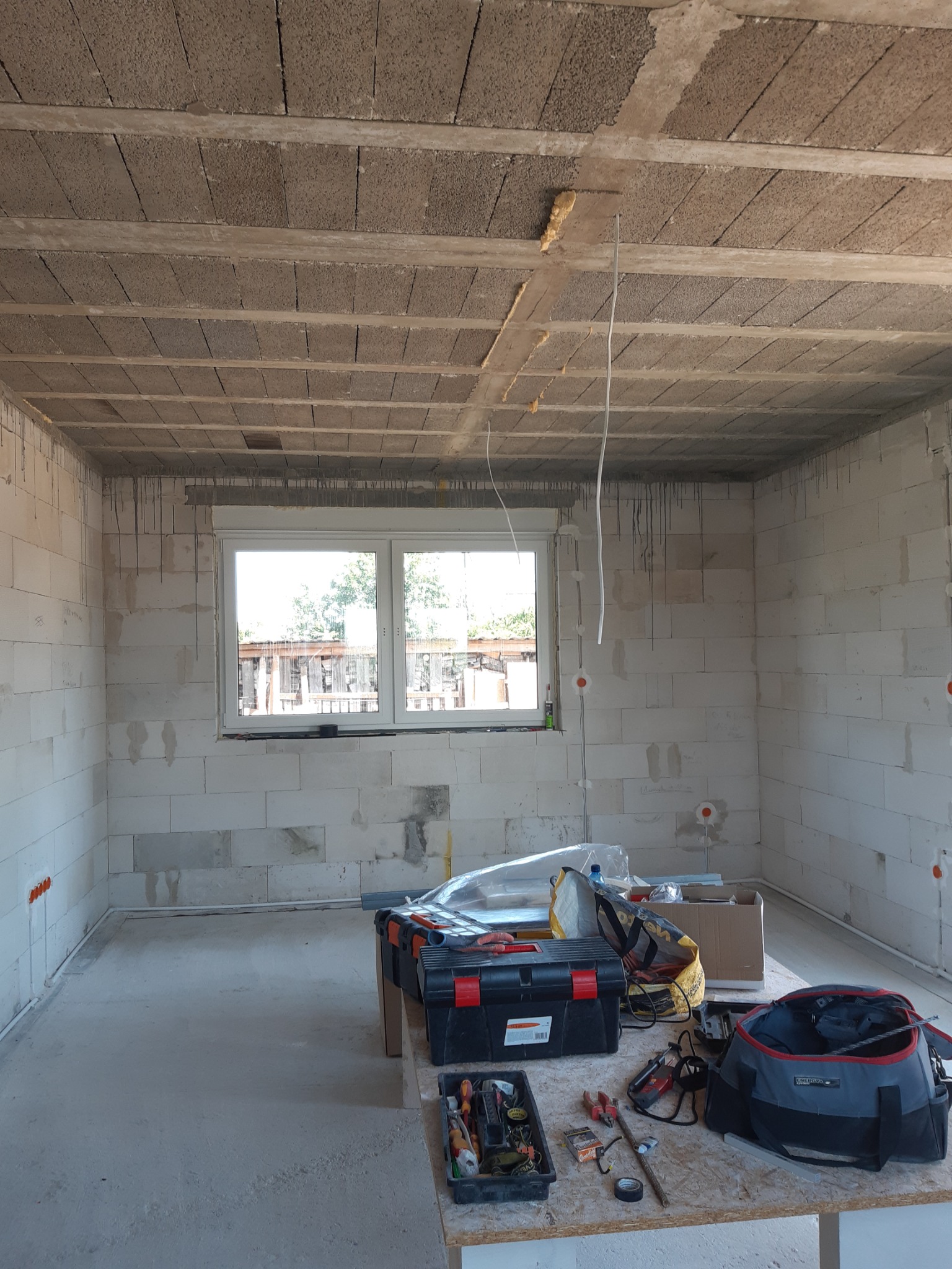 Surowy stan wnętrza z widocznymi przewodami elektrycznymi wystającymi z sufitu i ścian, skrzynkami narzędziowymi oraz torbą monterską na stole roboczym, okno w tle.