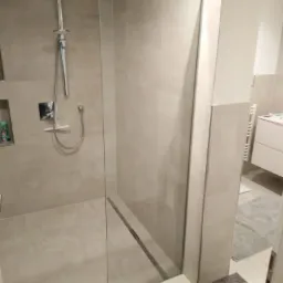 Beżowa kabina prysznicowa walk-in z odpływem liniowym, chromowaną armaturą i półką w ścianie, widok z zewnątrz na fragment łazienki z szafką umywalkową i dywanikiem.