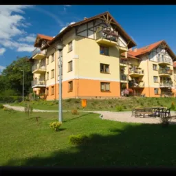 Nowoczesny, trzypiętrowy budynek apartamentowy z balkonami, w kolorach żółtym i pomarańczowym, z drewnianymi elementami dekoracyjnymi, otoczony zielonym trawnikiem i alejką z jasnego żwiru, na tle...