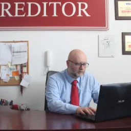 Mężczyzna w okularach i czerwonym krawacie pracuje na laptopie przy biurku. Na ścianie za nim wisi tablica z napisem 'CREDITOR' oraz dyplomy partnerskie Radia VOX FM.