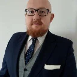 Portret mężczyzny w okularach, z rudą brodą, ubranego w elegancki, granatowy garnitur z szarą kamizelką i krawatem w kropki, stojącego na tle jasnej ściany.