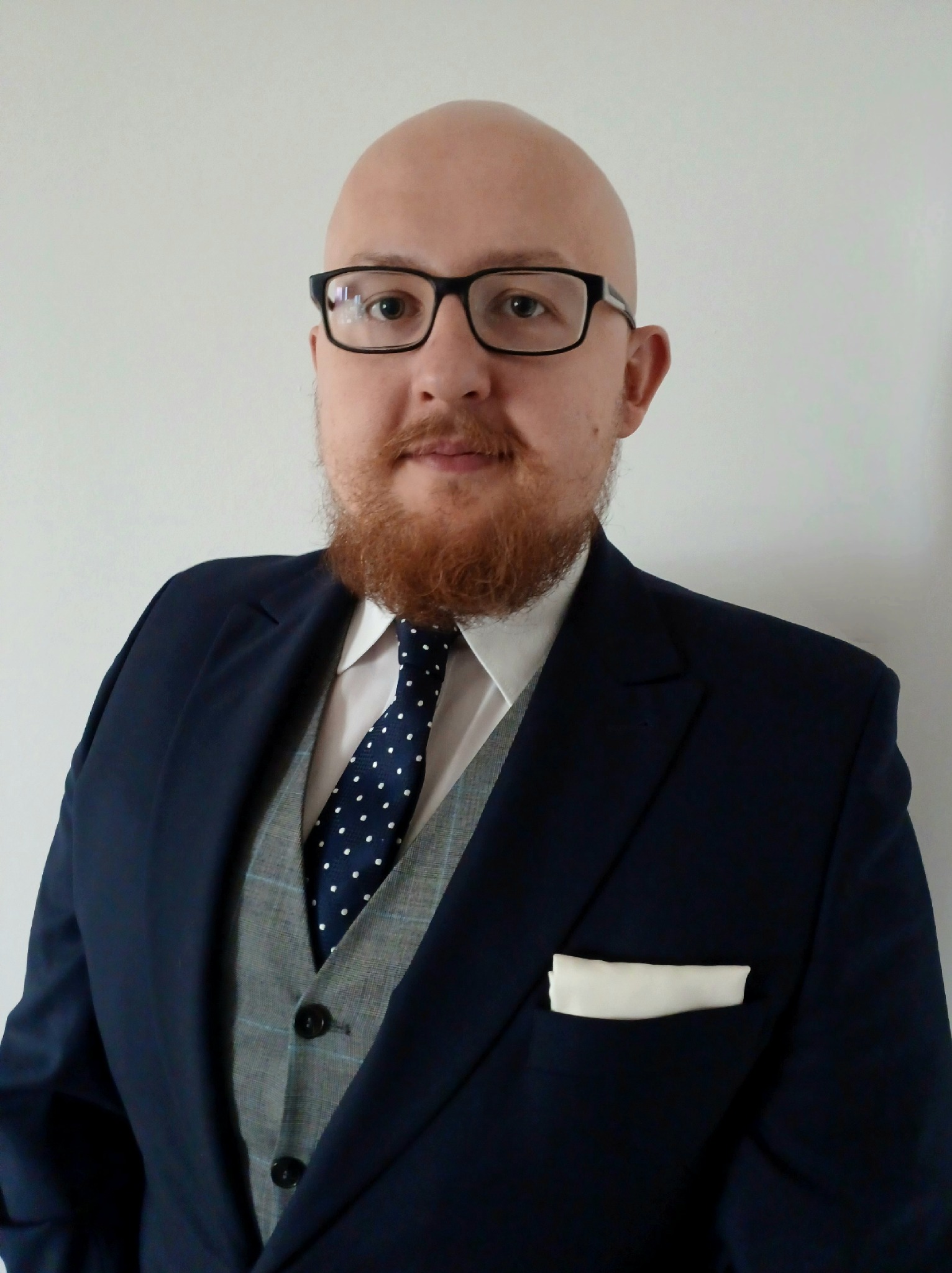 Portret mężczyzny w okularach, z rudą brodą, ubranego w elegancki, granatowy garnitur z szarą kamizelką i krawatem w kropki, stojącego na tle jasnej ściany.