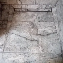 Wykończona kabina prysznicowa z szarymi płytkami imitującymi marmur i liniowym odpływem. Płytki ułożone w regularny wzór, z widocznymi fugami. Minimalistyczny design.