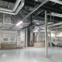 Wnętrze budynku komercyjnego z widoczną instalacją wentylacyjną, w tym izolowane kanały i okrągłe nawiewniki z czerwonymi elementami, z szarym betonowym podłożem.