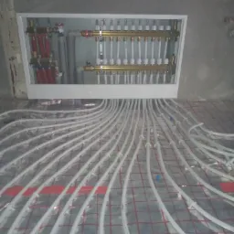 Rozdzielacz ogrzewania podłogowego w białej skrzynce, z licznymi białymi rurkami na folii z czerwoną siatką, podczas montażu w surowym pomieszczeniu.