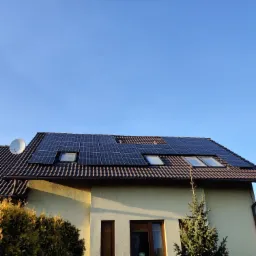 Panele fotowoltaiczne zamontowane na brązowym dachu domu jednorodzinnego z oknami dachowymi i anteną satelitarną, na tle błękitnego nieba.