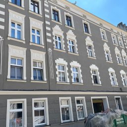 NOW BUD NOWAKOWSKI PAWEŁ - Szara kamienica po renowacji elewacji z dekoracyjnymi białymi obramowaniami okien. Kontener na gruz i częściowo widoczny samochód dostawczy.